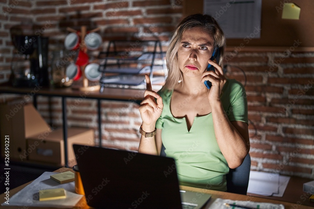 Young beautiful woman working at the office at night speaking on the phone pointing up looking sad and upset, indicating direction with fingers, unhappy and depressed. Young beautiful woman working at the office at night speaking on the phone pointing up looking sad and upset, indicating direction with fingers, unhappy and depressed.
