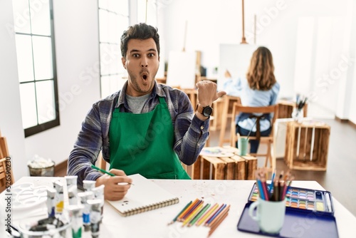 Young artist man at art studio surprised pointing with hand finger to the side, open mouth amazed expression.