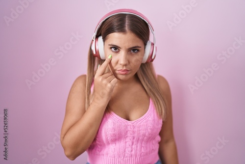 Young blonde woman listening to music using headphones pointing to the eye watching you gesture, suspicious expression