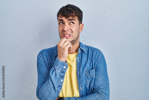 Young hispanic man standing over blue background thinking worried about a question, concerned and nervous with hand on chin