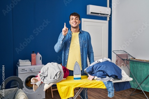 Young hispanic man ironing clothes at home smiling with an idea or question pointing finger up with happy face, number one