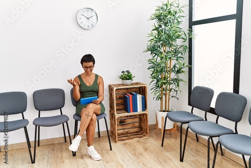 Young hispanic woman having video call sitting on chair at waiting room