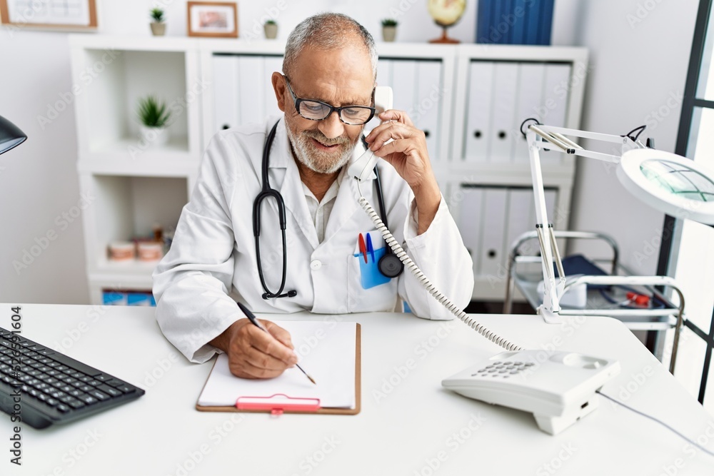 Senior grey-haired man wearing doctor uniform talking on the telephone at clinic Senior grey-haired man wearing doctor uniform talking on the telephone at clinic