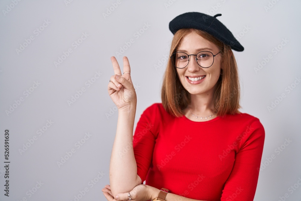 Young redhead woman standing wearing glasses and beret smiling with happy face winking at the camera doing victory sign with fingers. number two. Young redhead woman standing wearing glasses and beret smiling with happy face winking at the camera doing victory sign with fingers. number two.