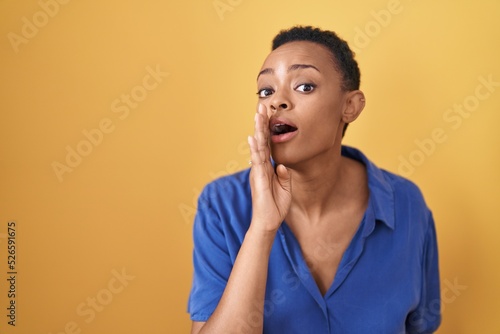African american woman standing over yellow background hand on mouth telling secret rumor, whispering malicious talk conversation
