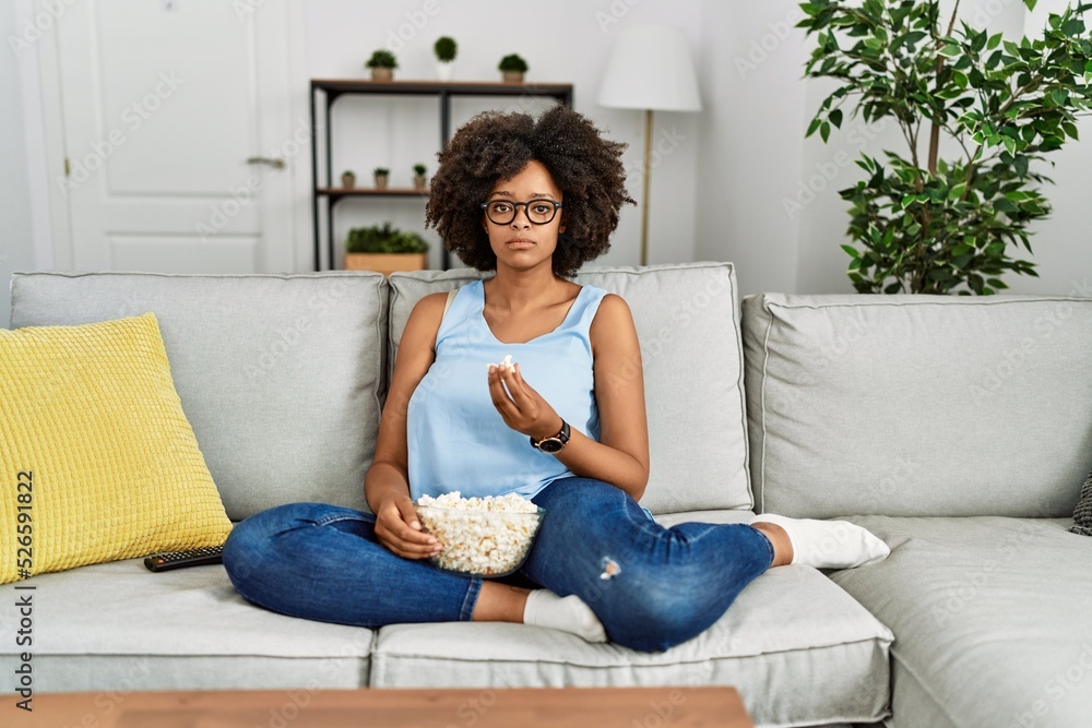 African american woman with afro hair sitting on the sofa eating popcorn at home thinking attitude and sober expression looking self confident African american woman with afro hair sitting on the sofa eating popcorn at home thinking attitude and sober expression looking self confident