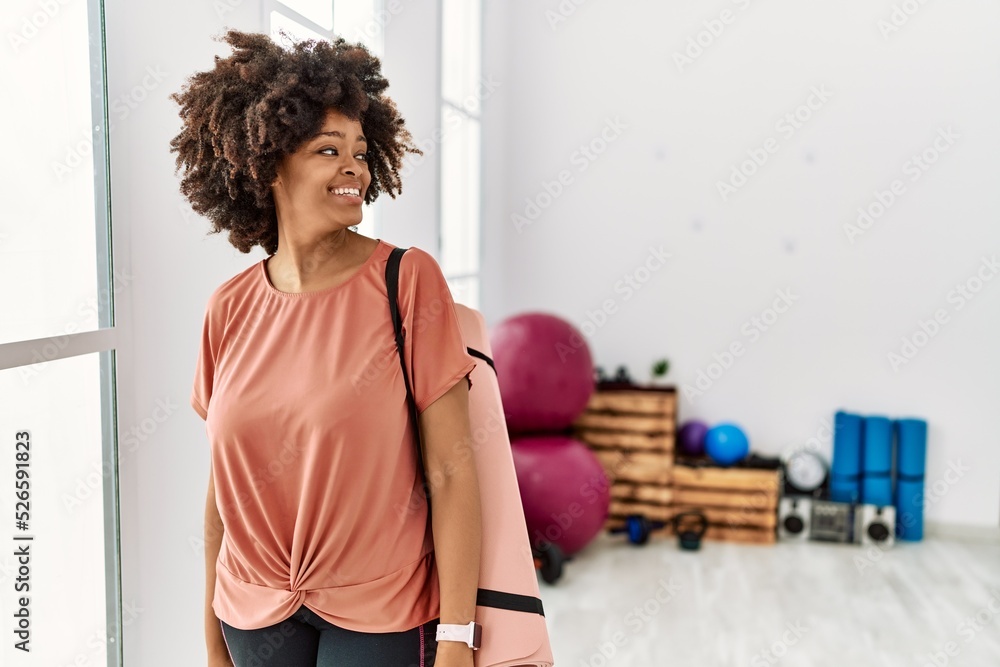 African american woman with afro hair holding yoga mat at pilates room looking away to side with smile on face, natural expression. laughing confident. African american woman with afro hair holding yoga mat at pilates room looking away to side with smile on face, natural expression. laughing confident.