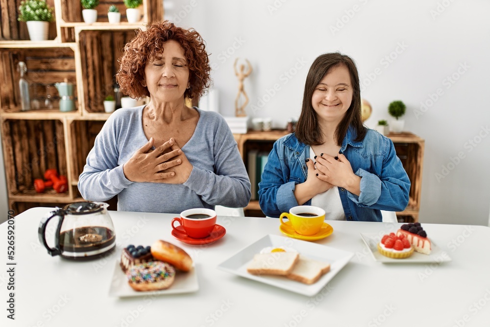Family of mother and down syndrome daughter sitting at home eating breakfast smiling with hands on chest with closed eyes and grateful gesture on face. health concept.