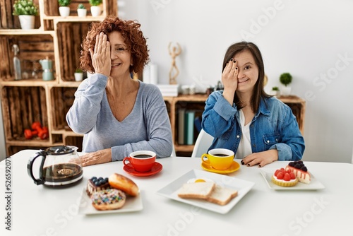 Family of mother and down syndrome daughter sitting at home eating breakfast covering one eye with hand  confident smile on face and surprise emotion.