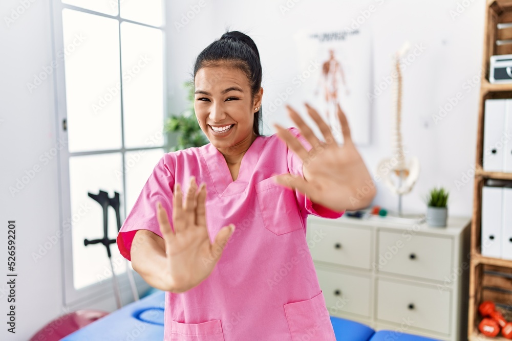 Young hispanic woman working at pain recovery clinic afraid and terrified with fear expression stop gesture with hands, shouting in shock. panic concept. Young hispanic woman working at pain recovery clinic afraid and terrified with fear expression stop gesture with hands, shouting in shock. panic concept.
