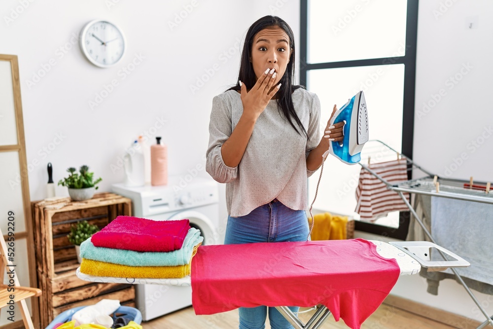 Young hispanic woman ironing clothes at home covering mouth with hand, shocked and afraid for mistake. surprised expression