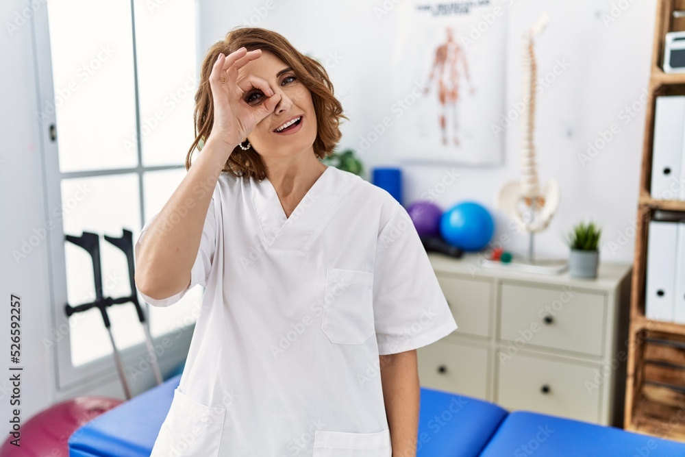 Middle age physiotherapist woman working at pain recovery clinic doing ok gesture with hand smiling, eye looking through fingers with happy face.