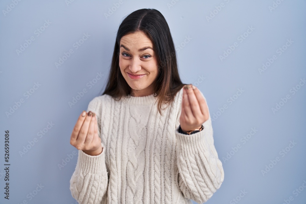 Young brunette woman standing over blue background doing money gesture with hands, asking for salary payment, millionaire business
