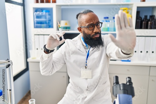 African american man working at scientist laboratory holding syringe with open hand doing stop sign with serious and confident expression  defense gesture