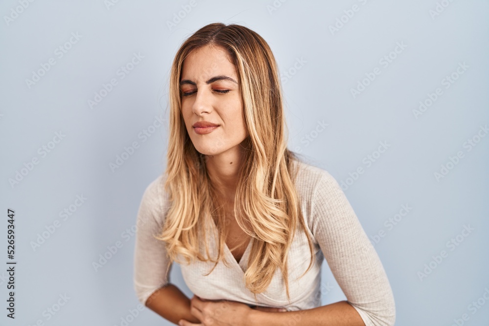Young blonde woman standing over isolated background with hand on stomach because indigestion, painful illness feeling unwell. ache concept. Young blonde woman standing over isolated background with hand on stomach because indigestion, painful illness feeling unwell. ache concept.