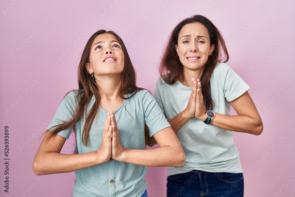 Young mother and daughter standing over pink background begging and praying with hands together with hope expression on face very emotional and worried. begging. Young mother and daughter standing over pink background begging and praying with hands together with hope expression on face very emotional and worried. begging.