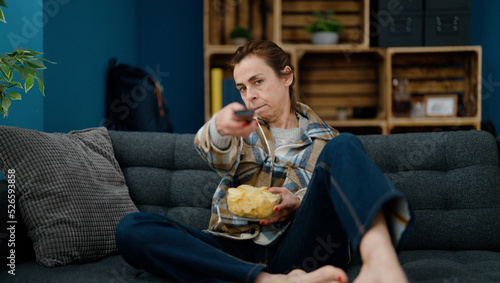 Middle age hispanic woman eating chips potatoes watching movie at home