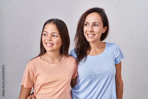 Young mother and daughter standing over white background looking away to side with smile on face, natural expression. laughing confident.