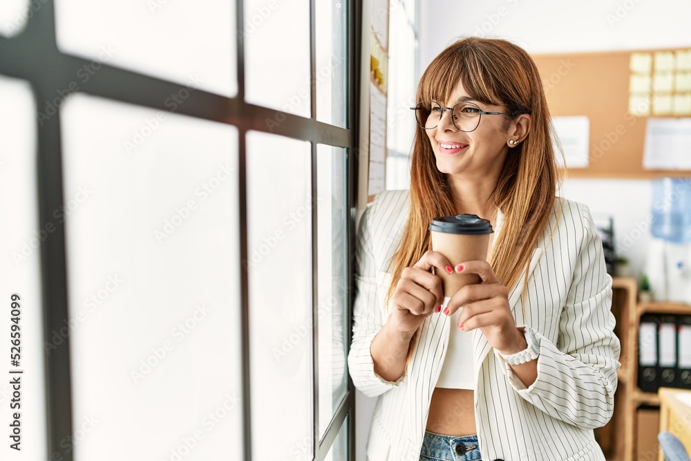 Young hispanic businesswoman smiling happy drinking coffee standing by the window at the office. Young hispanic businesswoman smiling happy drinking coffee standing by the window at the office.