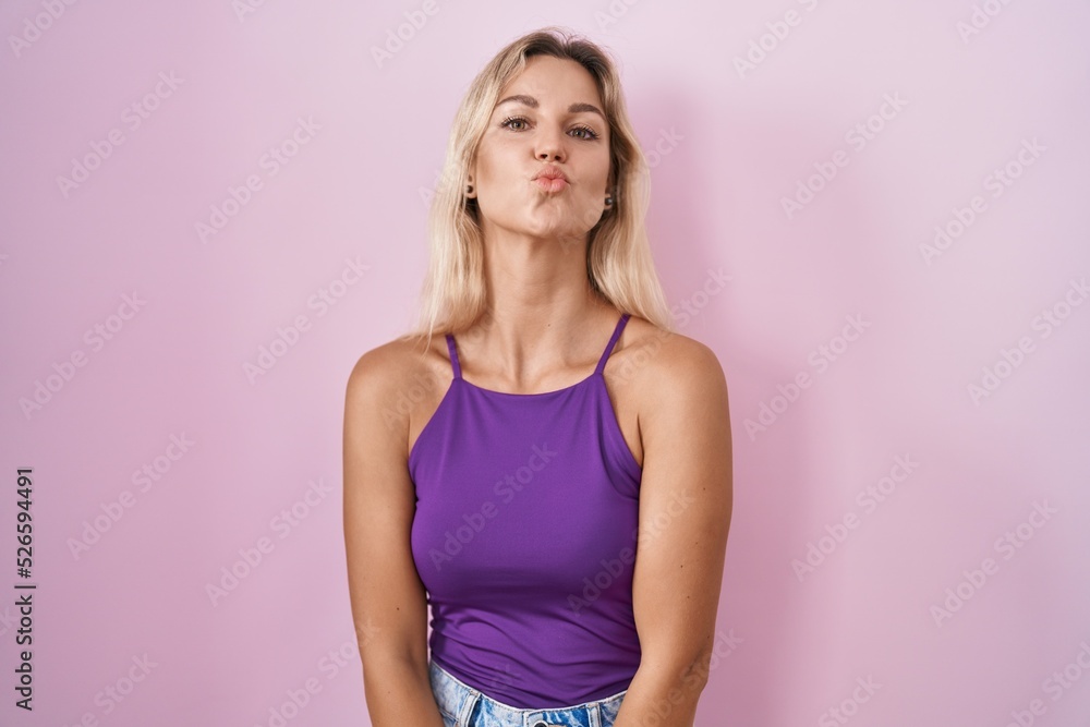 Young blonde woman standing over pink background looking at the camera blowing a kiss on air being lovely and sexy. love expression. Young blonde woman standing over pink background looking at the camera blowing a kiss on air being lovely and sexy. love expression.