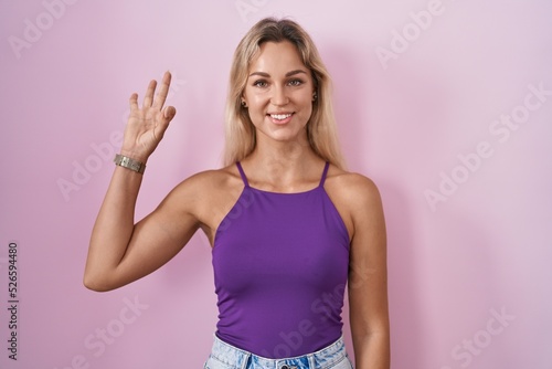Young blonde woman standing over pink background smiling positive doing ok sign with hand and fingers. successful expression.