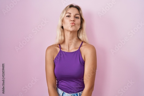 Young blonde woman standing over pink background looking at the camera blowing a kiss on air being lovely and sexy. love expression.