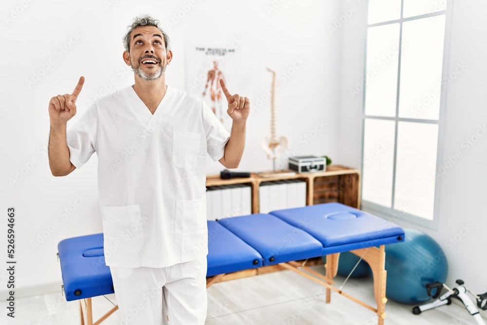 Middle age hispanic therapist man working at pain recovery clinic smiling amazed and surprised and pointing up with fingers and raised arms.