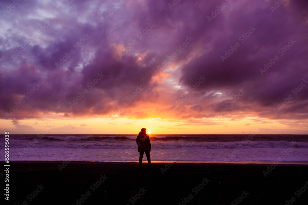 silueta de persona masculina parada en la orilla de la playa mirando al atardecer mientras el sol se oculta en el mar con nubes