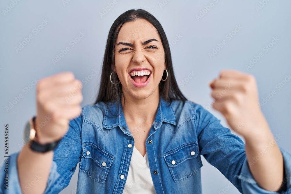 Hispanic woman standing over blue background angry and mad raising fists frustrated and furious while shouting with anger. rage and aggressive concept. Hispanic woman standing over blue background angry and mad raising fists frustrated and furious while shouting with anger. rage and aggressive concept.