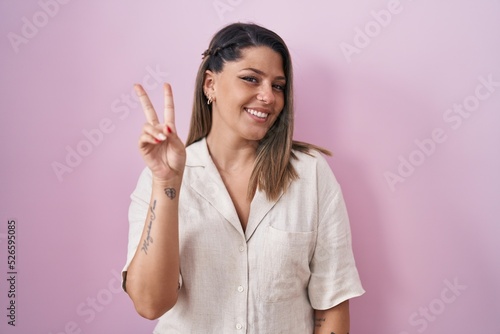 Blonde woman standing over pink background smiling looking to the camera showing fingers doing victory sign. number two.