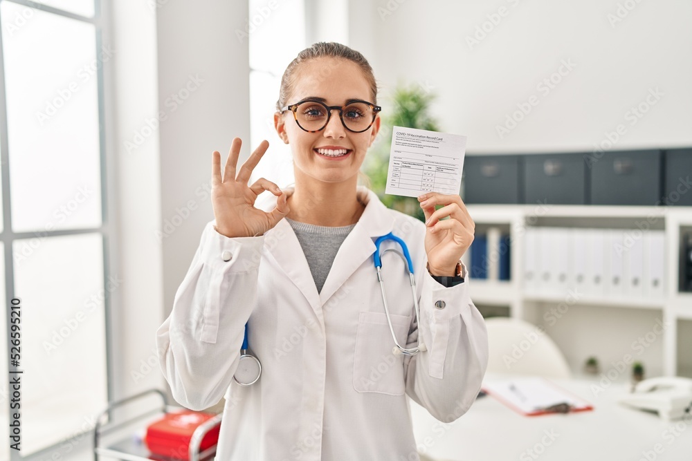 Young doctor woman holding covid certificate doing ok sign with fingers, smiling friendly gesturing excellent symbol