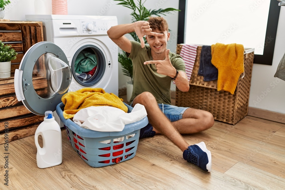 Young caucasian man putting dirty laundry into washing machine smiling making frame with hands and fingers with happy face. creativity and photography concept. Young caucasian man putting dirty laundry into washing machine smiling making frame with hands and fingers with happy face. creativity and photography concept.