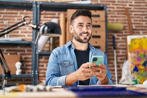 Young hispanic man using smartphone sitting on table at art studio