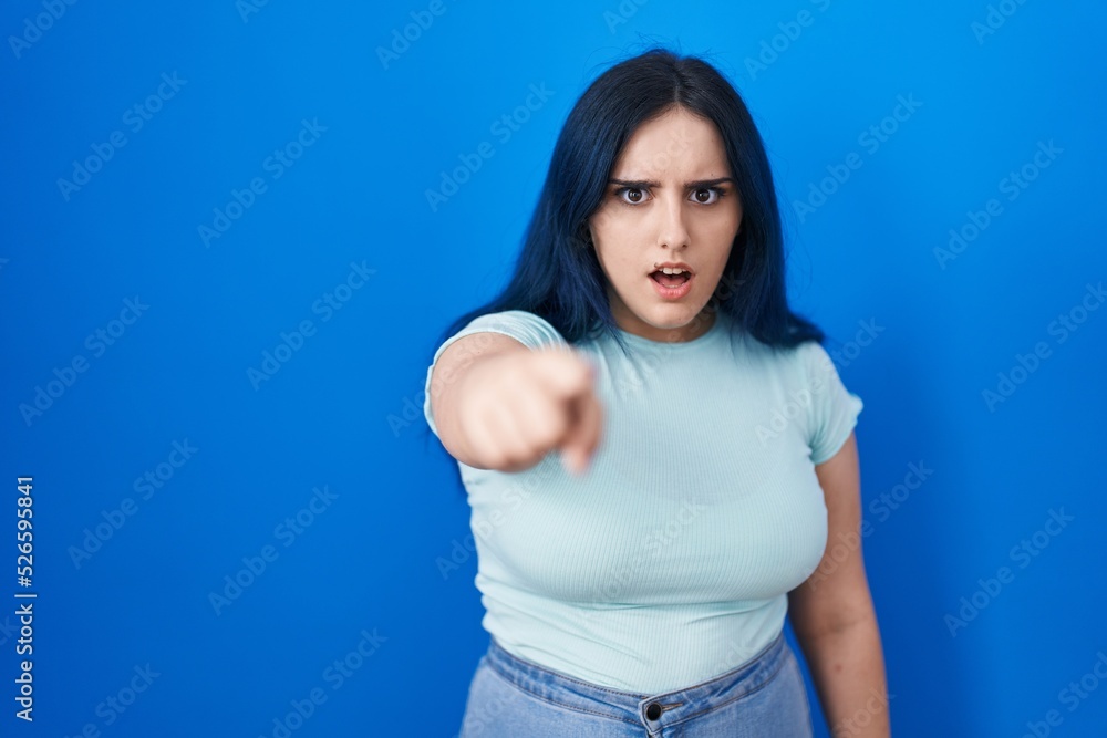 Young modern girl with blue hair standing over blue background pointing displeased and frustrated to the camera, angry and furious with you Young modern girl with blue hair standing over blue background pointing displeased and frustrated to the camera, angry and furious with you