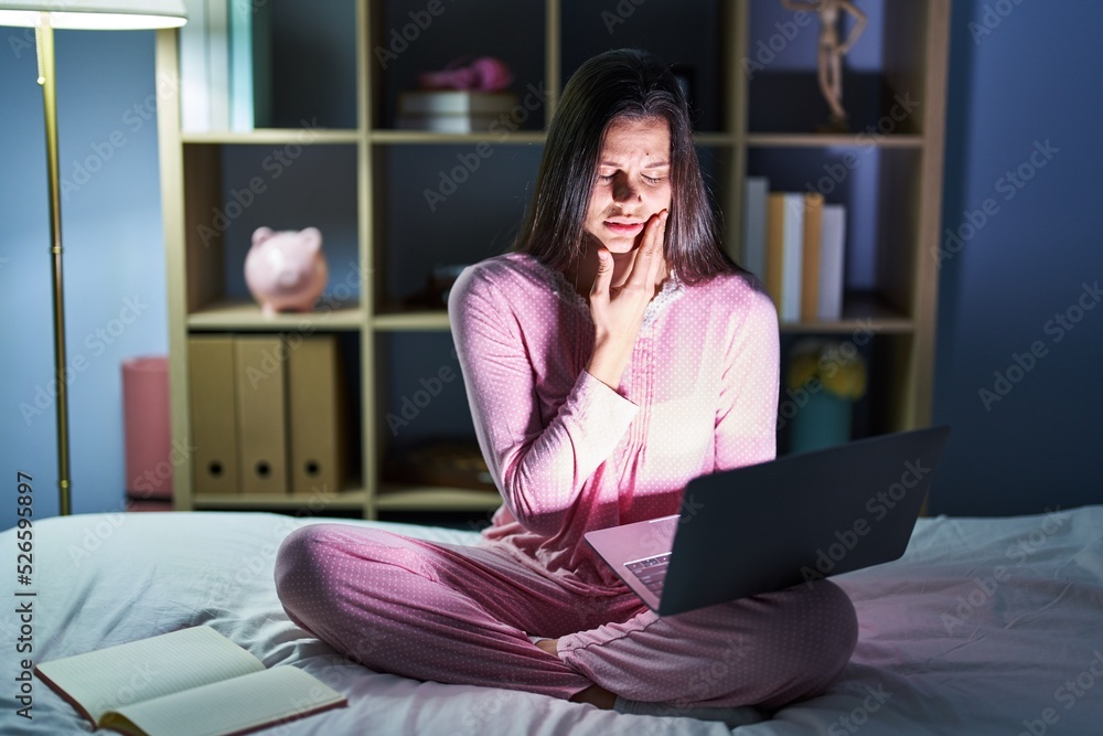 Young hispanic woman using computer laptop on the bed touching mouth with hand with painful expression because of toothache or dental illness on teeth. dentist concept. Young hispanic woman using computer laptop on the bed touching mouth with hand with painful expression because of toothache or dental illness on teeth. dentist concept.