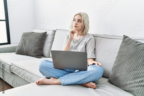 Young caucasian woman using laptop at home sitting on the sofa with hand on chin thinking about question, pensive expression. smiling with thoughtful face. doubt concept.