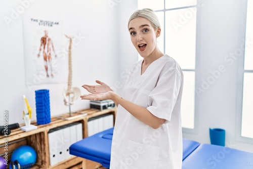 Young caucasian woman working at pain recovery clinic pointing aside with hands open palms showing copy space  presenting advertisement smiling excited happy