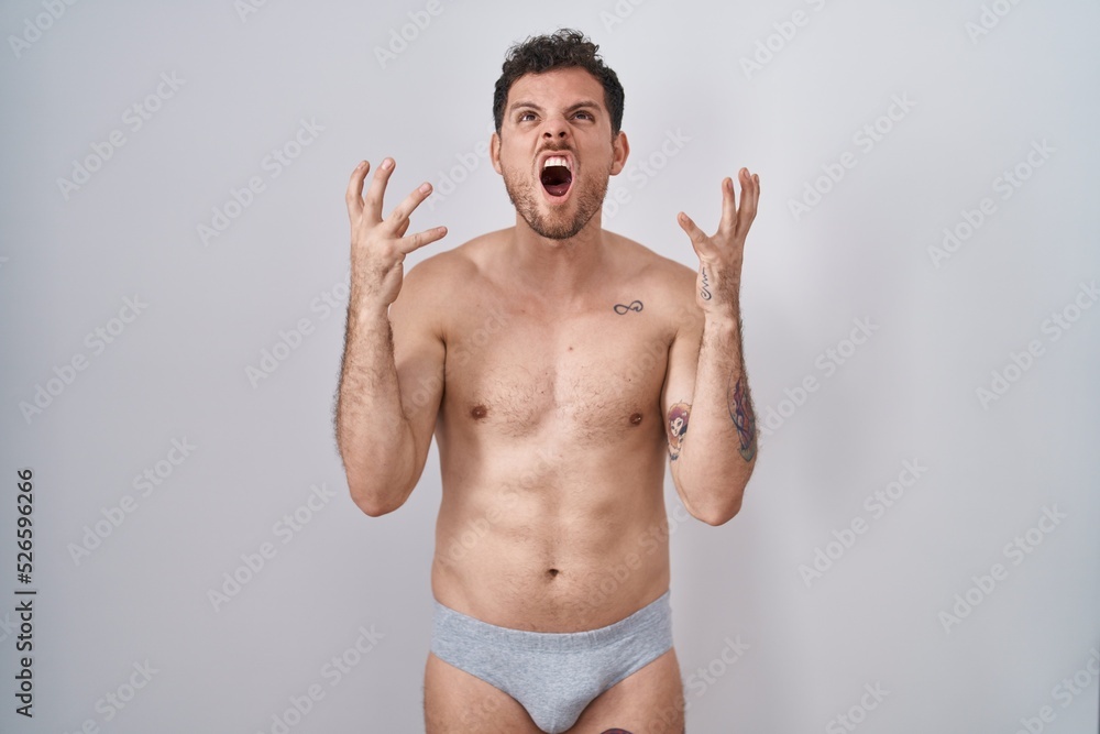 Fototapeta premium Young hispanic man standing shirtless wearing underware crazy and mad shouting and yelling with aggressive expression and arms raised. frustration concept.