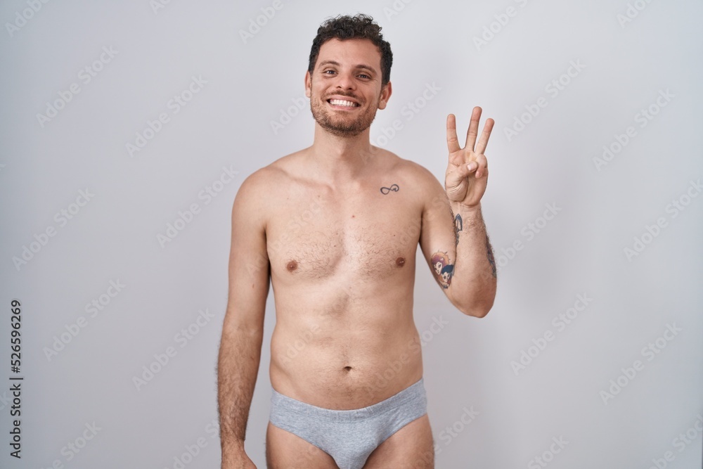 Young hispanic man standing shirtless wearing underware showing and pointing up with fingers number three while smiling confident and happy.