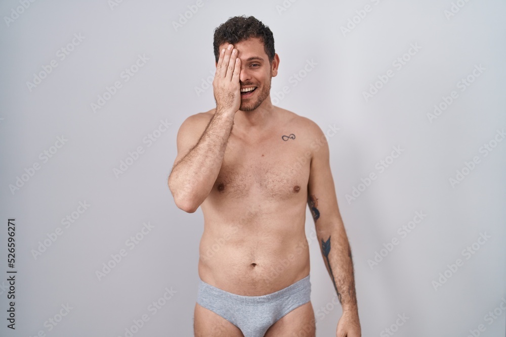Young hispanic man standing shirtless wearing underware covering one eye with hand, confident smile on face and surprise emotion.
