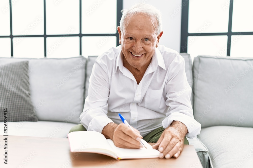 Senior man writing on book sitting on sofa at home Senior man writing on book sitting on sofa at home