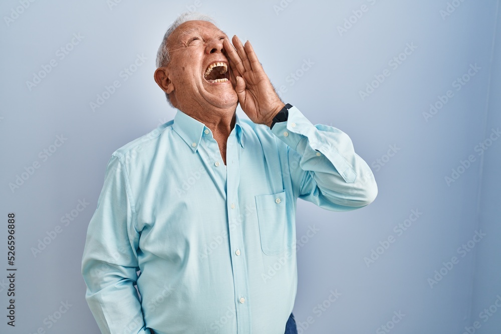 Senior man with grey hair standing over blue background shouting and screaming loud to side with hand on mouth. communication concept. Senior man with grey hair standing over blue background shouting and screaming loud to side with hand on mouth. communication concept.