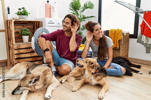 Photography Young hispanic couple doing laundry with dogs smiling with hand over ear listening an hearing to rumor or gossip