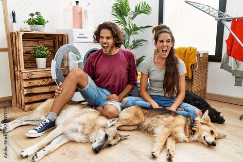 Young hispanic couple doing laundry with dogs angry and mad screaming frustrated and furious, shouting with anger. rage and aggressive concept.
