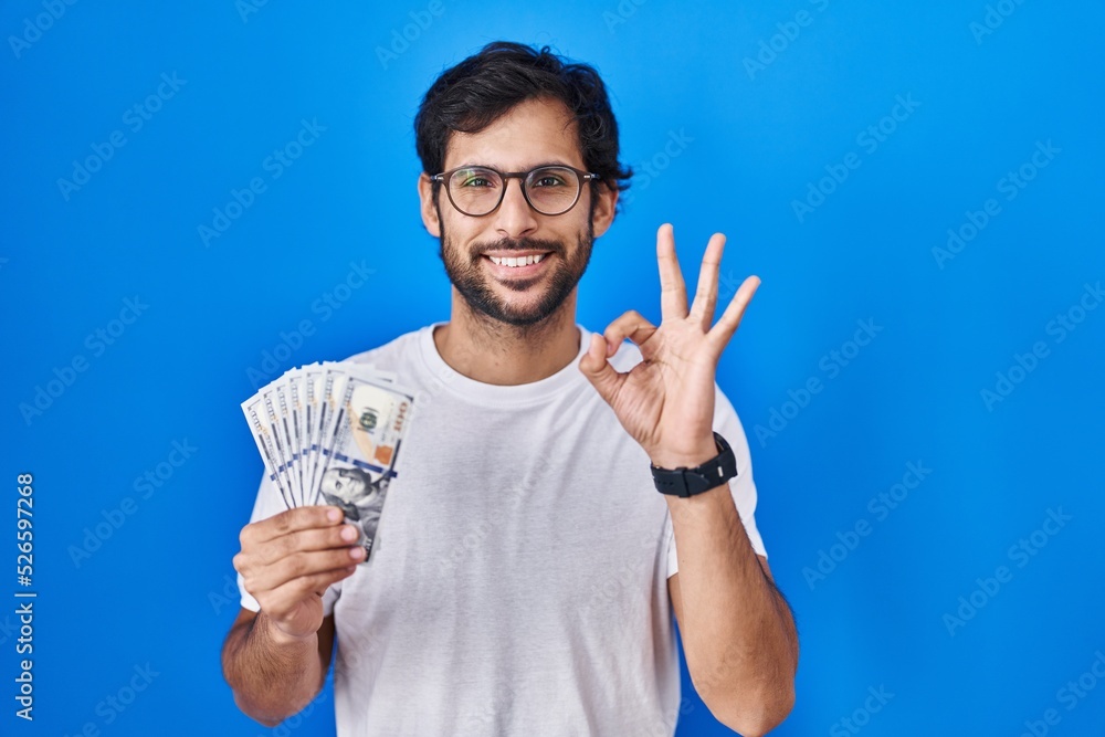 Naklejka premium Handsome latin man holding dollars banknotes doing ok sign with fingers, smiling friendly gesturing excellent symbol