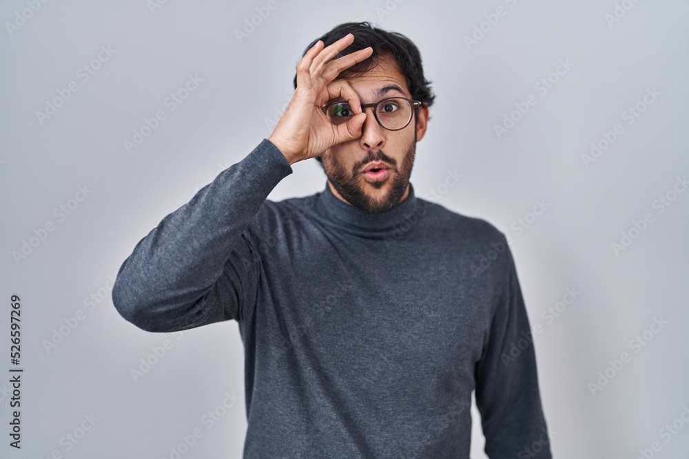 Handsome latin man standing over isolated background doing ok gesture shocked with surprised face, eye looking through fingers. unbelieving expression. Handsome latin man standing over isolated background doing ok gesture shocked with surprised face, eye looking through fingers. unbelieving expression.