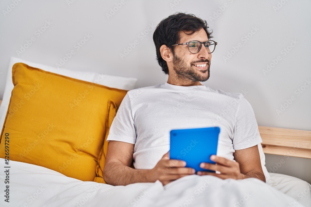 Young hispanic man using touchpad sitting on bed at bedroom Young hispanic man using touchpad sitting on bed at bedroom