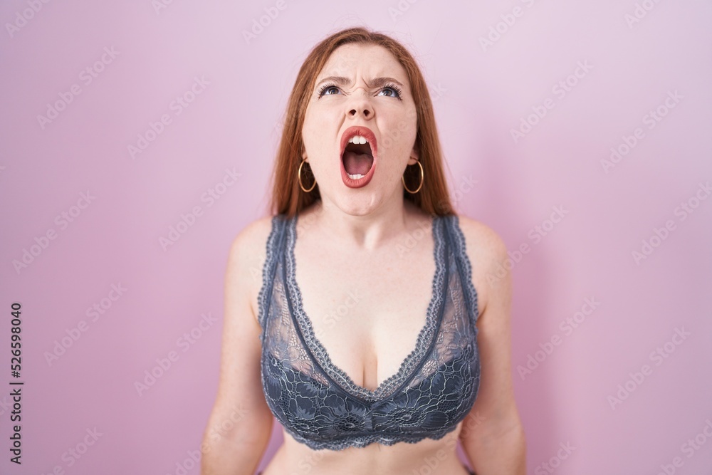 Redhead woman wearing lingerie over pink background angry and mad screaming frustrated and furious, shouting with anger. rage and aggressive concept. Redhead woman wearing lingerie over pink background angry and mad screaming frustrated and furious, shouting with anger. rage and aggressive concept.