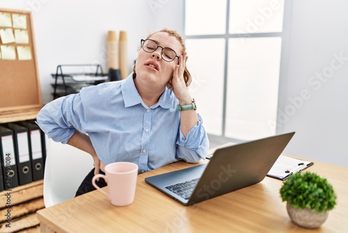 Young redhead woman working at the office using computer laptop stretching back, tired and relaxed, sleepy and yawning for early morning