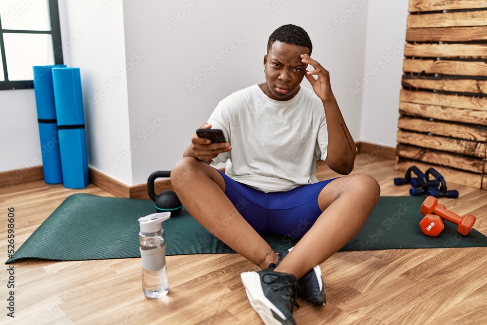 Obraz premium Young african man sitting on training mat at the gym using smartphone worried and stressed about a problem with hand on forehead, nervous and anxious for crisis